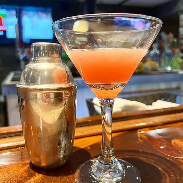 A pink cocktail in a martini glass and a silver shaker on a wooden bar at Yamas Bar & Grill.