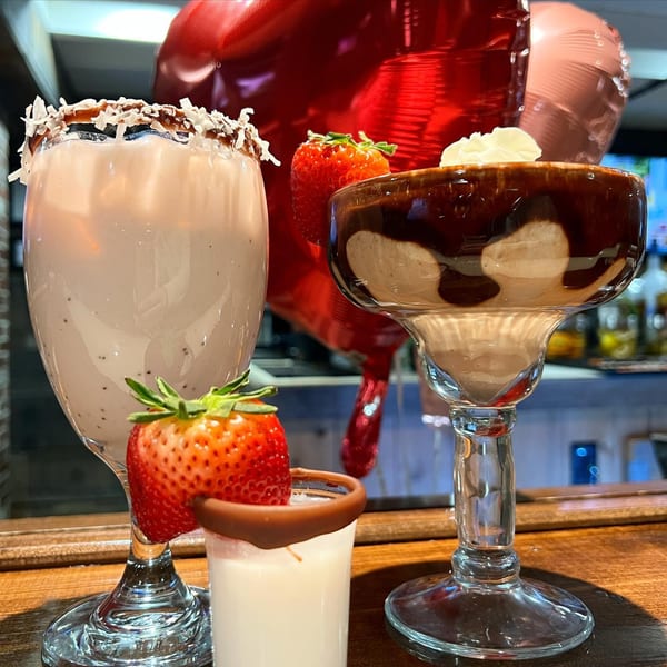 Two Greek dessert cocktails with strawberries and chocolate, one with coconut flakes, on a wooden bar.