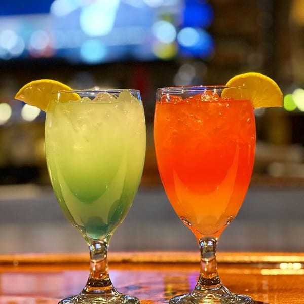 Two vibrant cocktails, one green and one orange, garnished with lemon slices on a bar.