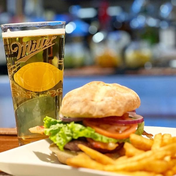A burger with lettuce, tomato, and onion, served with fries and a glass of Miller beer.